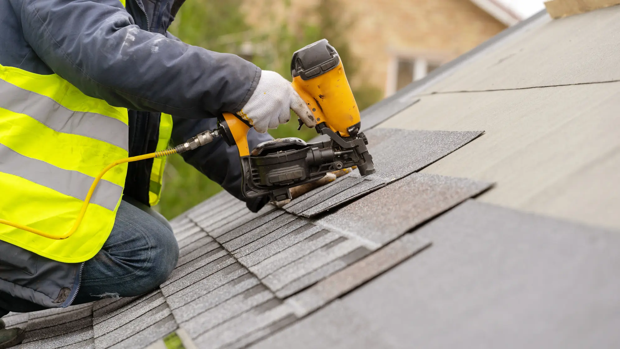Roofing contractor installing asphalt shingles on a residential roof using a nail gun.